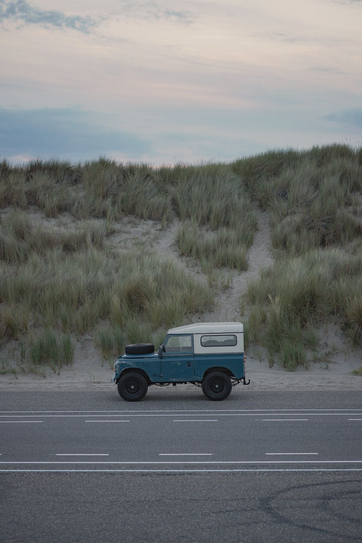A Defender parked roadside at dusk, dunes and sea grass beyond.